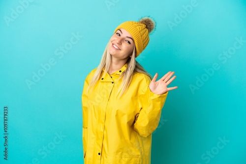 Young woman wearing a rainproof coat over isolated blue background saluting with hand with happy expression