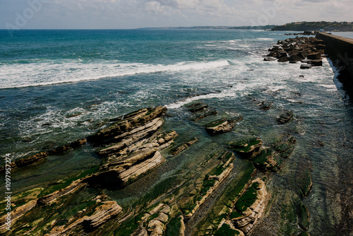 Rocky coastal landscape with green algae-covered rocks and clear blue ocean waves under a partly cloudy sky, creating a serene and natural seascape. photo