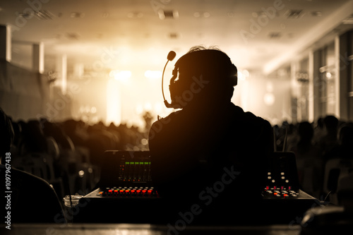 Silhouette of a sound engineer wearing a headset, managing audio equipment during a live performance. Warm golden light illuminates the stage and the crowd, creating a dramatic event scene.