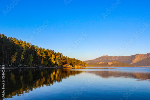 Autumn forest reflection in calm lake water with golden sunlight illuminating pine trees. Burabay, Kazakhstan