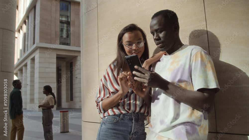 Two young people standing near urban building talking watching cellphone. African American and Latino student near campus communicating. Male and female coworkers on break chilling outdoors chatting. Two young people standing near urban building talking watching cellphone. African American and Latino student near campus communicating. Male and female coworkers on break chilling outdoors chatting.