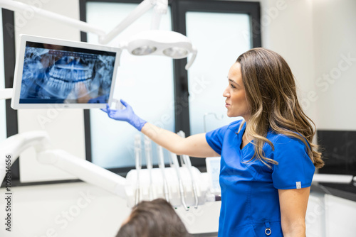 A female dentist in blue scrubs stands in a clinic, pointing at X-rays on a screen, as she explains the dental condition to an attentive patient in a chair. photo