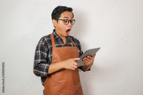 asian man wearing apron holding tablet with surprised expression looking at tablet, coffee and restaurant business idea concept photo