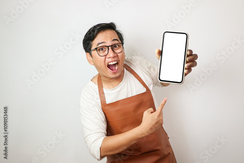 Shocked young asian man wearing rolled-up white sleeve shirt and brown apron is holding a smartphone with surprised face expression. Isolated over white background.