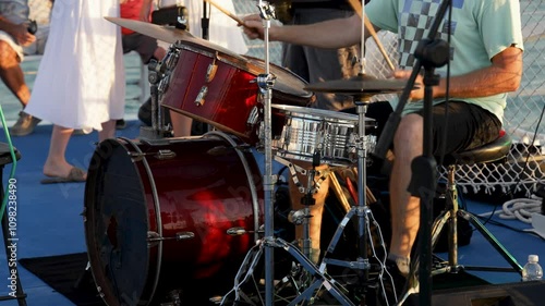 Close-up of a drummer playing a red drum set outdoors, surrounded by casually dressed people in a lively setting.