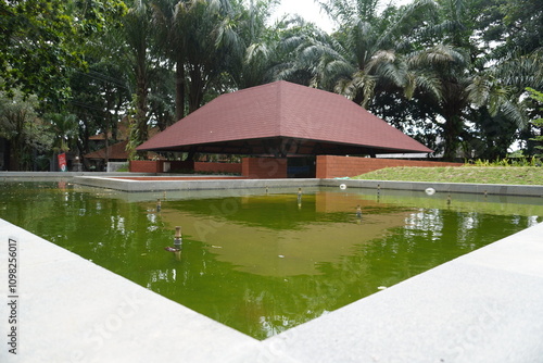 A semi-outdoor building with a pool in the middle and a red triangular roof which is a garden in a public space. photo