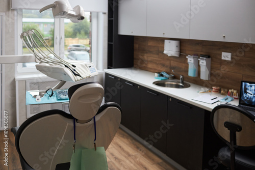 Modern dental clinic interior showing dental chair, equipment, and tools neatly organized. Room features large window providing natural lighting and wooden accents contributing to warm atmosphere photo