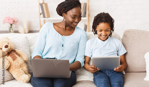 Stay at home african mom working remotely on laptop and helping her child with homework, using tablet and laptop photo