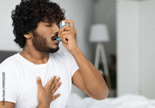 Indian guy suffering from asthma, using inhaler in bed, side view, copy space. Young hindu man woke up with asthma attack, touching chest and using nebulizer, bedroom interior