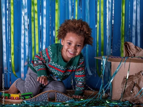 Obraz na plátne Smiling Multi ethnic boy at Christmas, wearing a festive jumper sits with his presents