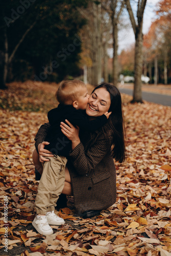A small blond son in a dark sweater and light pants stands on a leafy road and gently kisses his dark-haired mother in a checkered coat.