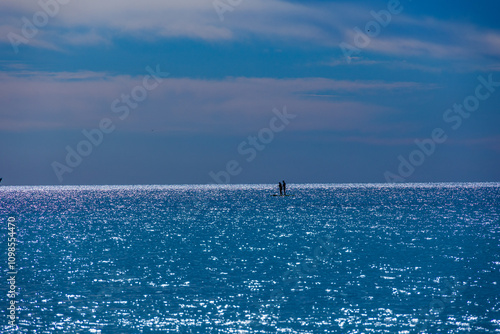 Sunlight shimmers across a vast expanse of calm water. Two silhouettes, possibly paddleboarders, photo