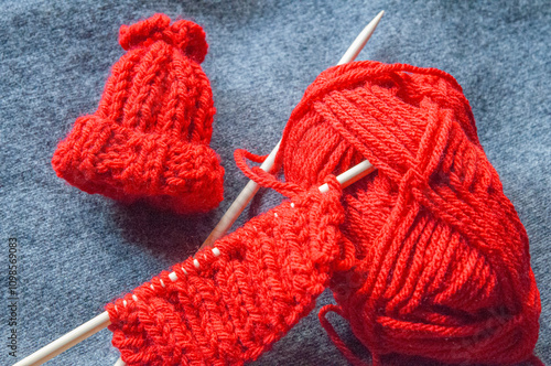 A red mini knit cap and knitting needles with an unfinished project. The scene captures a cozy, handmade crafting vibe, perfect for showcasing knitting, creativity, and DIY hobbies