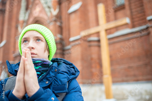 12 year old boy in green hat and blue jacket prays in front of catholic church