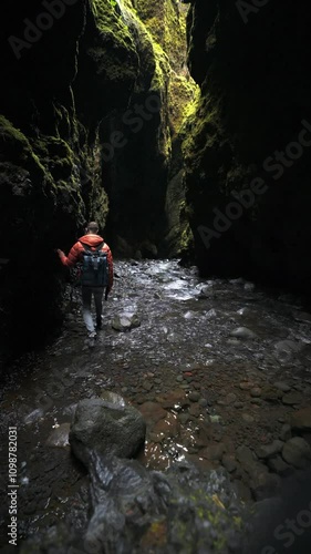 A person in an orange jacket walks through a narrow canyon in Iceland, with steep walls, green moss, and a shallow stream creating a unique landscape.
