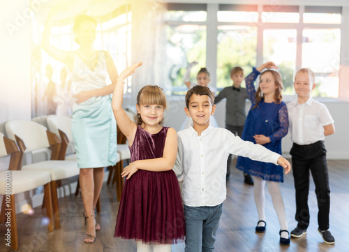 Happy interested preteen pupils preparing for festive school event, rehearsing ballroom dance steps in spacious classroom while female teacher observing from background.. photo