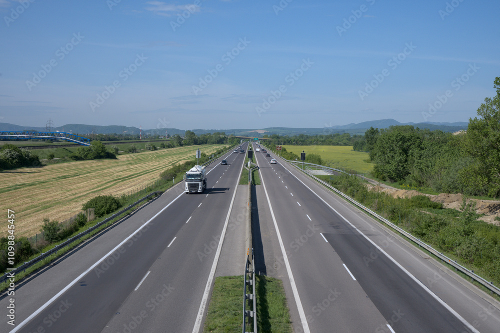 Fototapeta premium A highway stretching into the horizon, surrounded by green fields and trees under a clear blue sky.