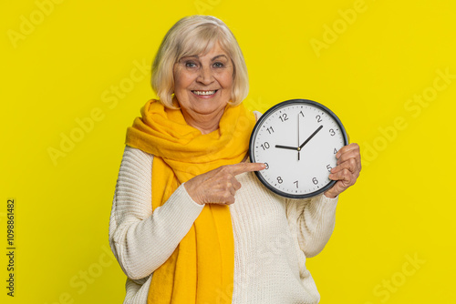 It is your time, hurry up. Senior old woman showing time on wall office clock, ok thumb up approve pointing finger at camera, advertisement, deadline. Elderly grandmother isolated on yellow background photo