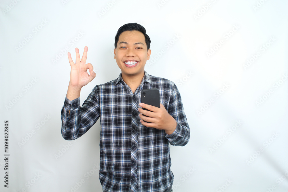 Fototapeta premium confident asian man smiling show okay symbols with finger and holding mobile phone isolated on white