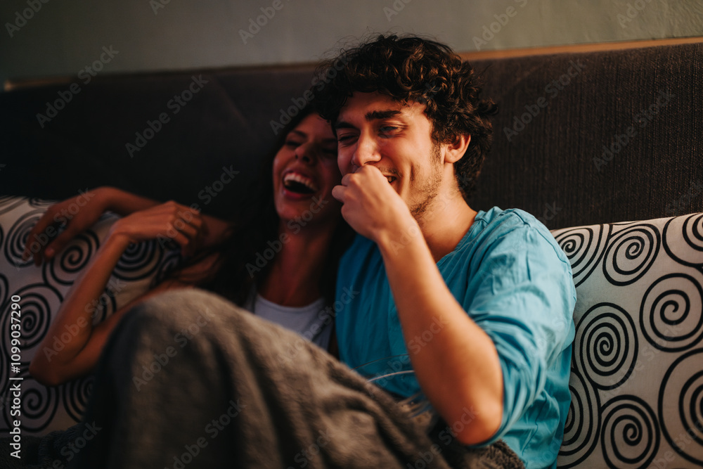 Fototapeta premium A joyful young couple shares a laugh while sitting on a cozy sofa covered in patterned pillows. Their close bond and happiness are evident, creating a warm and inviting atmosphere.