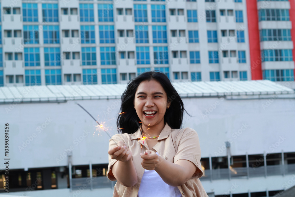 Fototapeta premium carefree asian young woman holding sparkler celebrate new year eve with laugh and dance in rooftop apartment outdoor with city building background