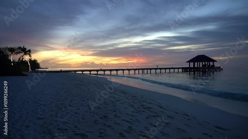 Wallpaper Mural An orange sunset on a tropical island with white sand. Green palm trees, trees and bushes. Gray clouds are highlighted in orange-red. The calm ocean is like a mirror. A bridge and a house on the water Torontodigital.ca