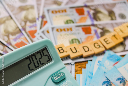 Calculator with 2025 displayed, banknotes, budgeting tiles, an alarm clock, eyeglasses, a game controller, and a cup of tea on a table. New year finance, money and budget concept