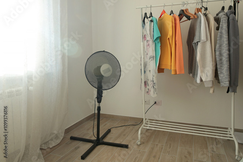 Fan on floor of room near window and clothes hangers. Coolness and seasons. Heat and summer.  photo