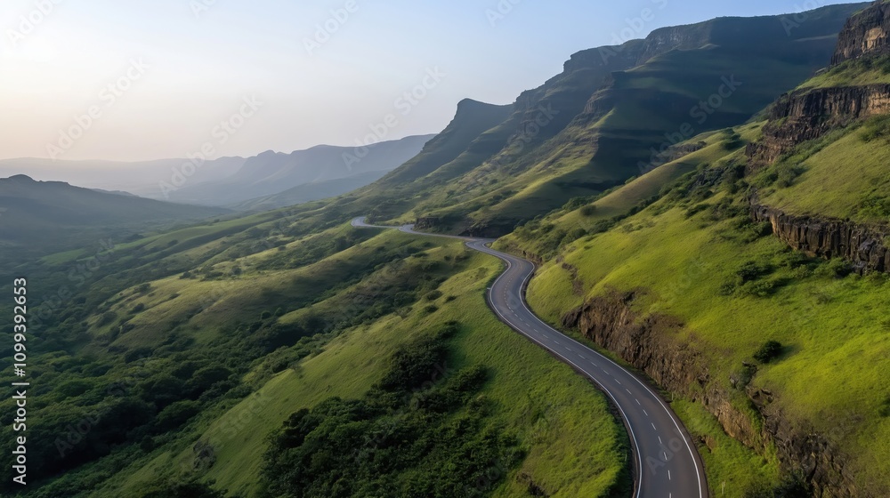Fototapeta premium Winding road through a lush green mountainous landscape with rocky cliffs and a clear sky in the background