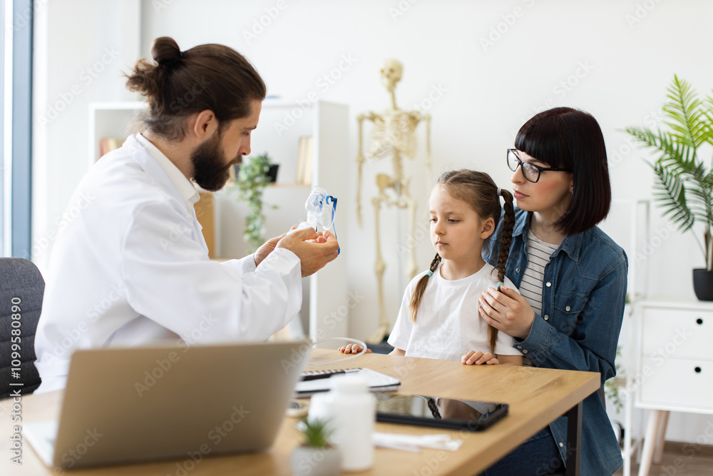 Fototapeta premium Caucasian male doctor offers nebulizer treatment to young Caucasian girl at clinic. Caring adult female, presumably mother, supports child during medical consultation.