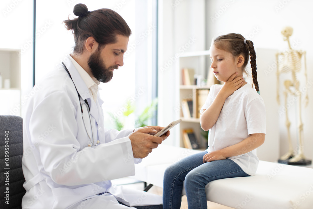 Fototapeta premium Caucasian male doctor with beard examines Caucasian young girl complaining of sore throat in medical office. Doctor checks symptoms using digital device while girl holds throat.