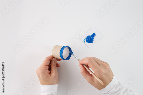 a person painting the edge of a cardboard tube with blue paint using a paintbrush. step in a process of crafting or DIY activity, creative use of everyday materials like a cardboard tube and paint photo