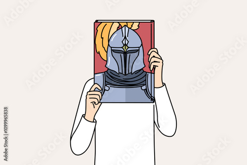 Book with knight on cover in hands of man covering face, reading literature about medieval soldiers. Man pretends to be knight in armor, dreaming of participating in gladiatorial tournaments.