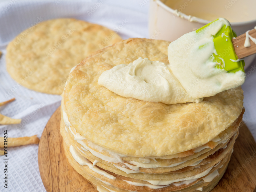 Silicone spatula spreading vanilla custard over a pile of baked thin round buttery layers on a wooden board, bowl and a pile waiting in background ready to make shortcrust puff pastry Napoleon cake