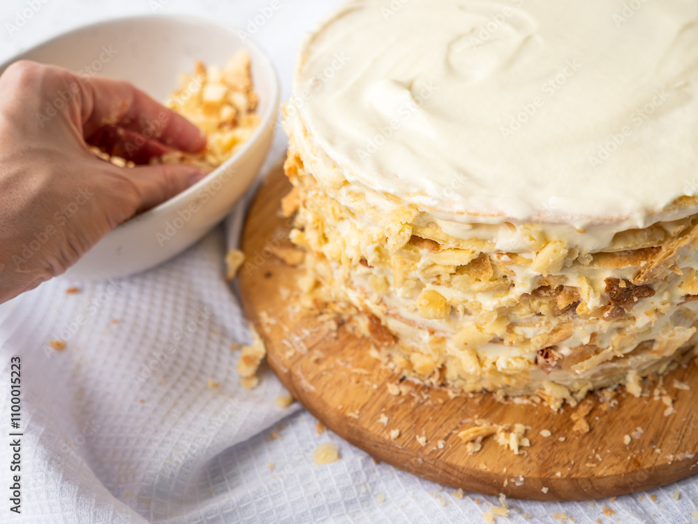 Hand sprinkling decorating crumbs over a pile of thin round layers spread with vanilla custard, bowl of dough crumbs on a white waffle towel in the blurry background. Shortcrust puff pastry Napoleon