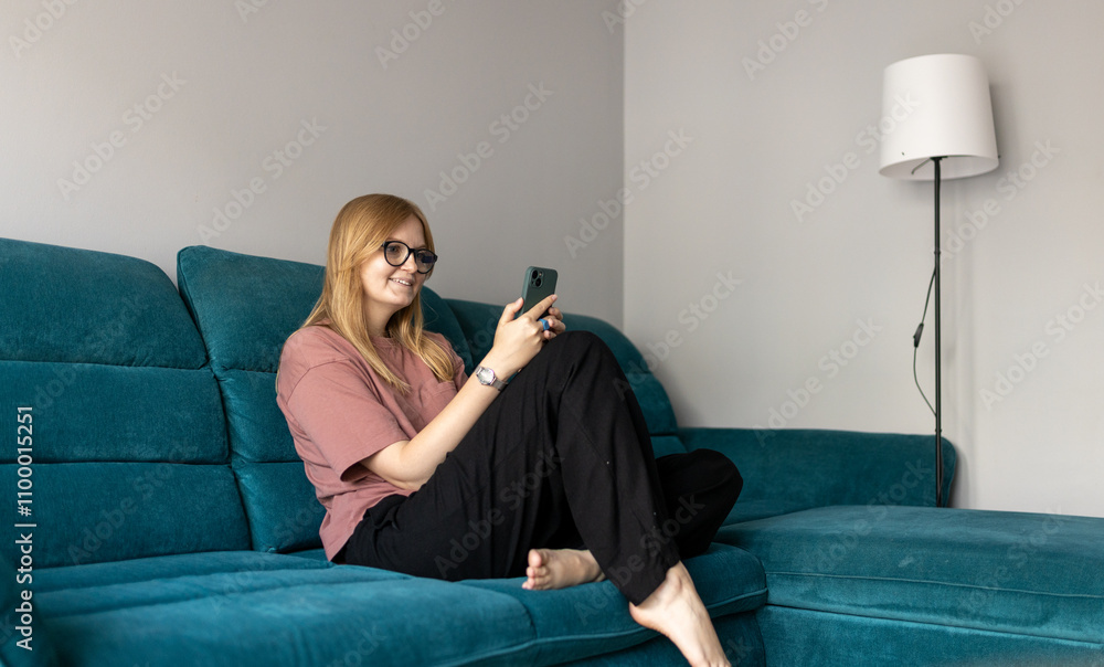 Obraz premium Beautiful blonde woman having fun online on her smartphone at home or hotel. Phone Communication and comfort concept. Smiling 30s woman daydreaming and holding mobile phone with copy space. Full body