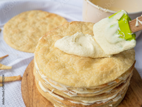 Obraz na płótnie Silicone spatula spreading vanilla custard over a pile of baked thin round buttery layers on a wooden board, bowl and a pile waiting in background ready to make shortcrust puff pastry Napoleon cake w salonie