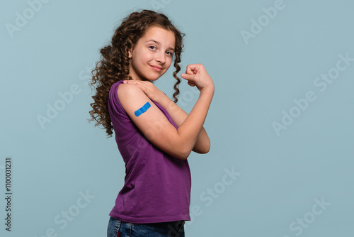 Girl flexing her arm with a blue bandage after receiving a vaccine shot, wearing a purple shirt