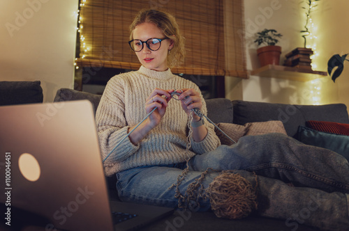 Young beautiful blonde woman with glasses learning to knit online sitting at home during Christmas holidays