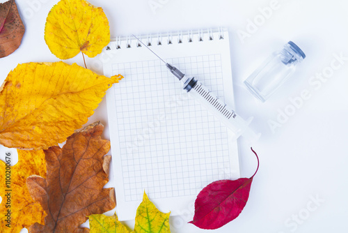 Vaccine and syringe are on table next to doctor's notebook and autumn leaves. Blank notebook for notes in doctor's office. Autumn vaccination..