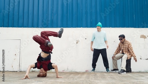 Young street dancer dancing in hip hop style with multicultural friends cheering behind at wall. Attractive handsome man stretch arms at street with blue background. Outdoor sport 2024. Hiphop. photo