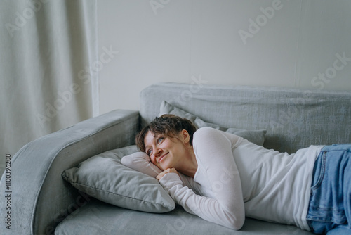 Young woman relaxes on a grey sofa at home, enjoying peace and quiet, embracing tranquility in casual wear with natural light