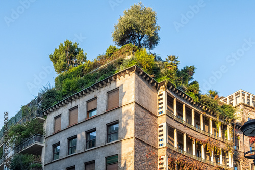 Urban building with lush rooftop garden, featuring trees and greenery cascading over the structure, blending architecture with nature under clear blue sky, Milan, Italy. Eco friendly urban environment