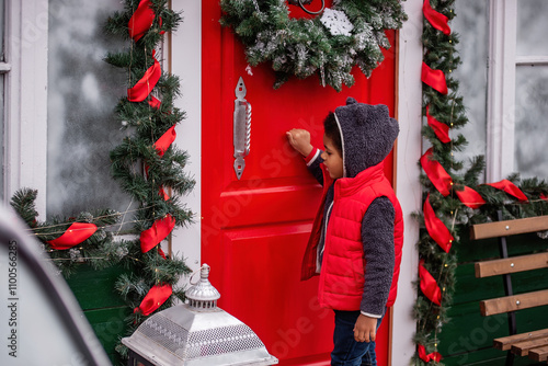 Cheerful boy in a cozy bear hoodie and red vest knocks on a festive red door adorned with holiday wreaths and garlands, capturing the magic of the Christmas season.