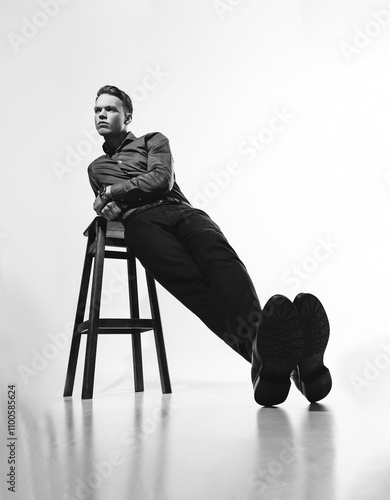 A stylish black and white portrait of a man in a shirt posing on a white cyclorama with a chair, evoking a sense of sophistication and refinement.