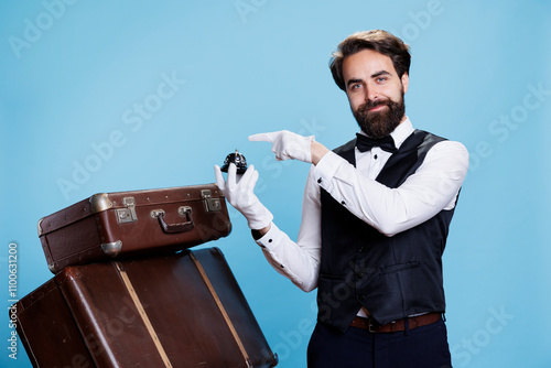 Hotel worker points to service bell, advertising hotel concierge assistance and ringing for help with luggage. Young formal doorkeeper helping with trolley bags, luxury profession. photo