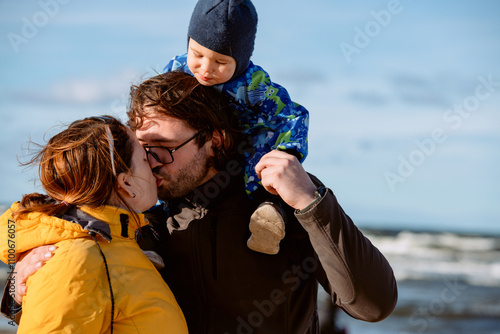 A happy family strolls along a chilly autumn beach, with their young son joyfully running ahead, creating lasting memories as the crisp wind and soothing waves embrace them in a moment of pure photo