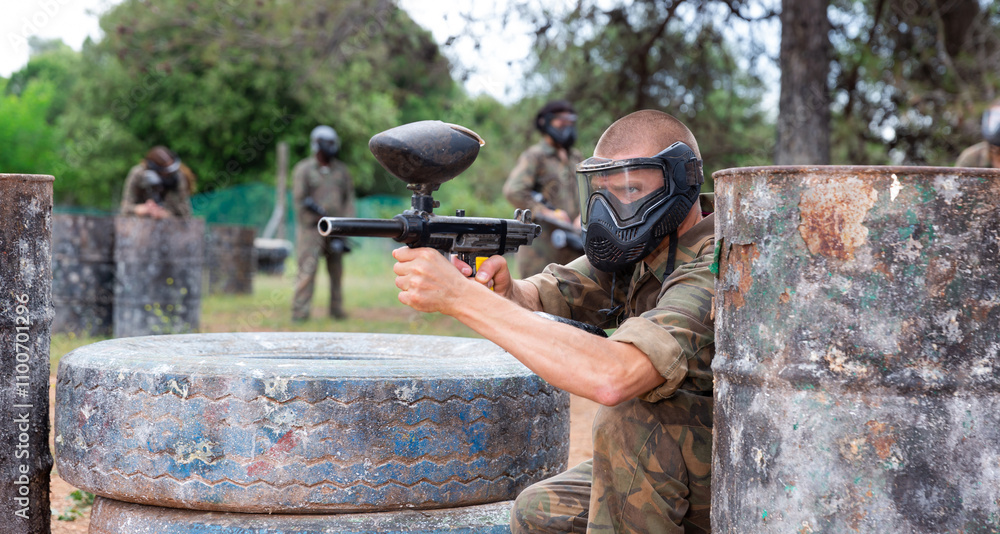 Fototapeta premium Paintball player in camouflage and mask aiming with gun in shootout outdoors