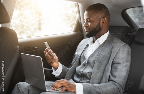 Handsome african american businessman with laptop holding smartphone while going by car on business trip, copy space. Black entrepreneur looking at mobile phone screen, using laptop photo