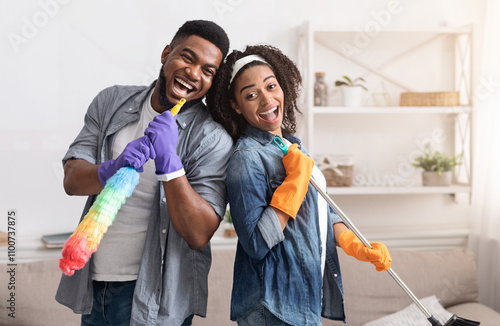 Domestic Band. Joyful Black Couple Having Fun During Spring-Cleaning Home, Singing Together, Using Mop Handle And Duster As Microphones